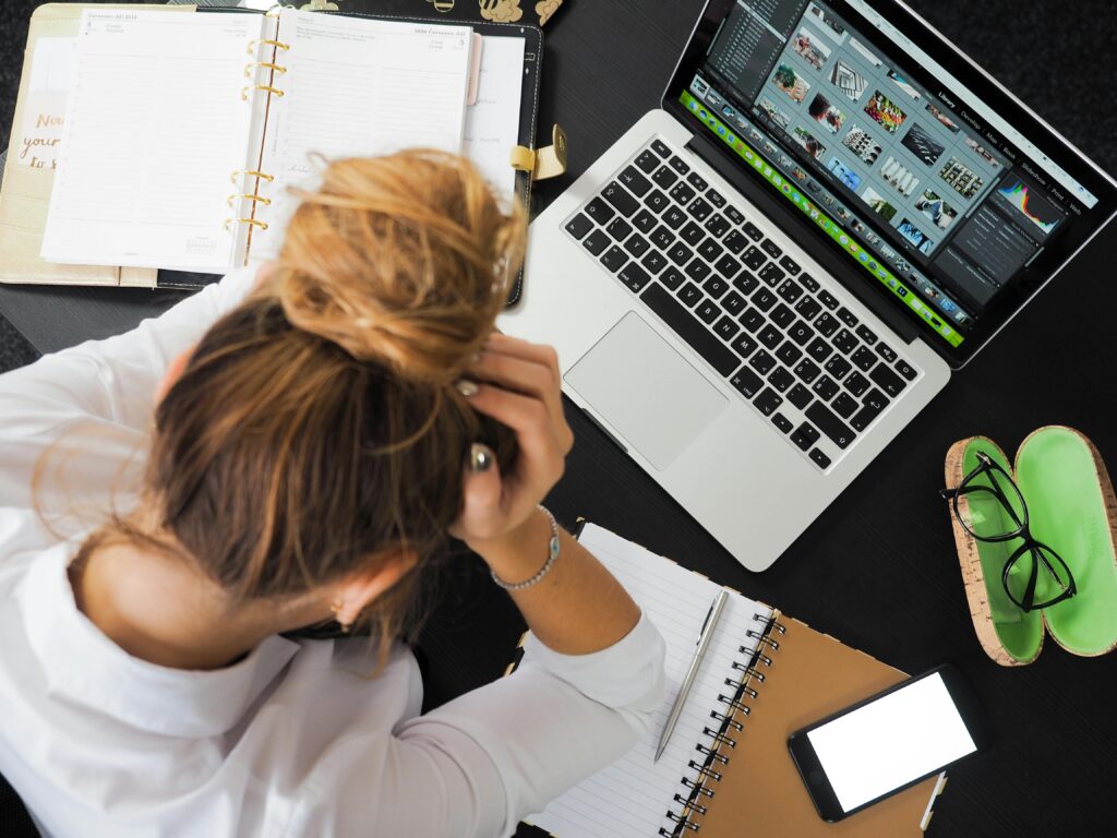 Woman sits in front of an open laptop, planner, and notebook with her head in her hands